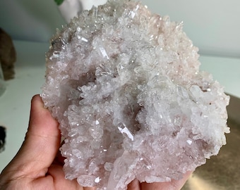 May include: A cluster of quartz crystals in shades of white and pale pink. The crystals are densely packed, creating a textured, spiky appearance. The quartz is held in a hand.
