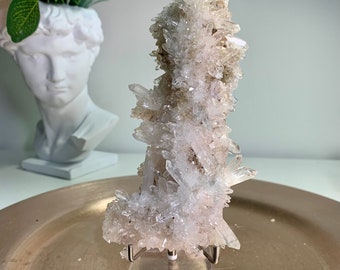 May include: A cluster of clear quartz crystals with a pale pink hue, mounted on a clear acrylic stand. The quartz formation has multiple points and a rough texture, displayed on a gold-toned metal tray.