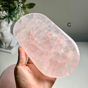 Pode incluir: Uma bandeja oval de quartzo rosa com uma superfície polida e translúcida, apresentando fraturas internas naturais e um tom rosa pálido. A bandeja está marcada com a letra C no canto superior direito e possui um design liso e plano.
