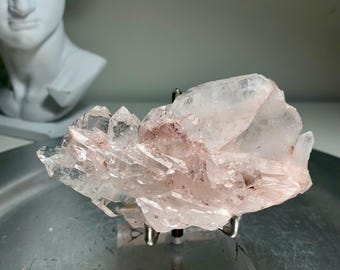 May include: A cluster of clear quartz crystals with reddish-brown inclusions, mounted on a metal stand. The crystals have sharp, pointed terminations and a translucent appearance. The quartz is displayed against a neutral background.