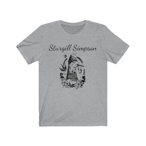 Pode incluir: Uma camiseta cinza mesclada com uma ilustração em preto e branco de um pássaro usando um chapéu e segurando um cachimbo. O texto "Sturgill Simpson" é impresso acima da ilustração.