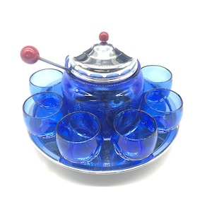 May include: A cobalt blue glass tea set with a silver-colored lid and tray. The set includes a teapot, six small glasses, and a stirring spoon. The teapot has a red knob on top and a red ball on the spoon handle.