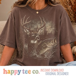 Puede incluir: Una camiseta marrón de manga corta con un estampado gráfico de un ciervo con grandes astas de pie en el agua junto a un pez saltando. El fondo del gráfico representa una escena forestal con árboles y hierba alta en tonos tierra.