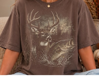 May include: A brown short-sleeve t-shirt featuring a graphic print of a deer with large antlers standing in water next to a jumping fish, with a forest background. The shirt has a crew neckline and a relaxed fit, showcasing a detailed illustration.