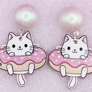 May include: A pair of dangle earrings featuring a white cat with pink and white donut. The donut has pink frosting and white sprinkles. The earrings have a pearl-like top.