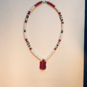 Peut inclure: Un collier de perles blanches et rouges avec une perle à motif noir et blanc. Le collier a un pendentif rouge en forme de bouclier.