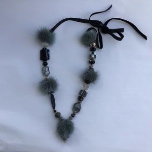 May include: A necklace with grey pom-poms, black beads, and a black ribbon tie. The necklace features a variety of shapes and textures, including fluffy pom-poms and geometric beads. The necklace is a unique accessory.