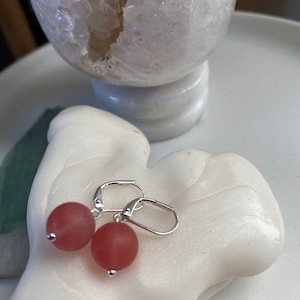 May include: Leverback earrings featuring matte, round, red beads. The earrings are presented on a white, sculpted hand-shaped object. A clear, spherical geode is in the background.