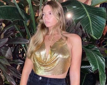 May include: A woman wearing a gold chainmail halter top with a deep V-neckline. The top is made of small, interconnected metal rings. She is standing in front of a large green plant.