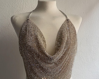 May include: Gold-colored chainmail halter top with a plunging neckline. The top is displayed on a mannequin against a white wall. The chainmail is made of small, reflective metal links.
