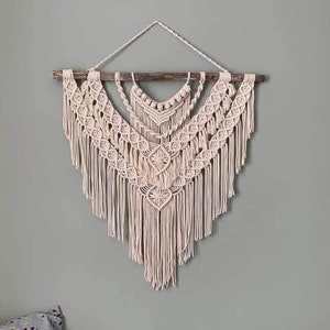 Peut inclure: Suspension murale en macramé crème avec un motif géométrique. La pièce est suspendue à une cheville en bois naturel et présente des motifs de nœuds complexes et de longues franges. Un textile décoratif pour la décoration intérieure.