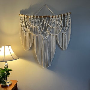 Puede incluir: Un gran tapiz de macram&eacute; blanco roto. La pieza est&aacute; suspendida de una rama de madera natural y presenta intrincados patrones anudados y largas y fluidas flecos. Un elemento decorativo para el hogar.