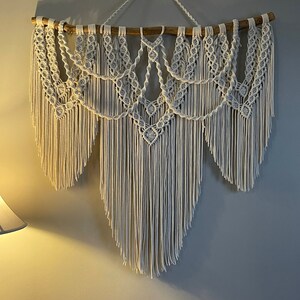 Puede incluir: Un gran tapiz de macram&eacute; blanco roto. La pieza presenta intrincados patrones de nudos y largas y fluidas franjas. Est&aacute; suspendido de una rama de madera natural, creando una est&eacute;tica bohemia y texturizada. Decoraci&oacute;n de pared.