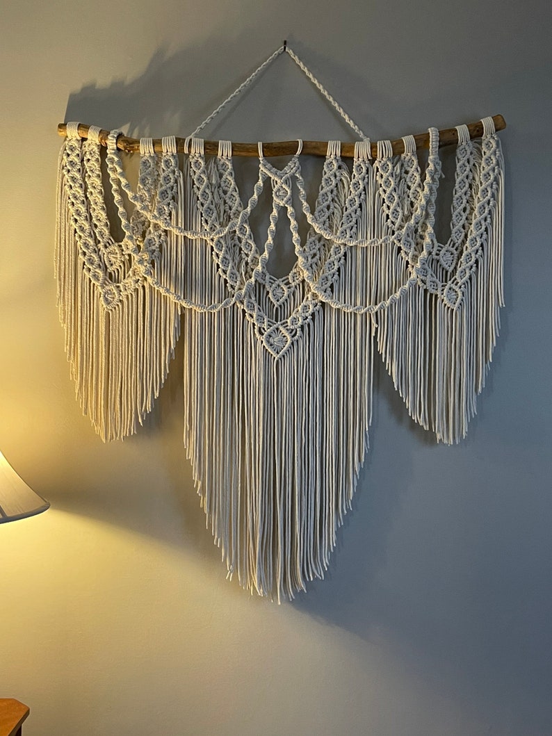 Puede incluir: Un gran tapiz de macram&eacute; blanco roto. La pieza presenta intrincados patrones de nudos y largas y fluidas franjas. Est&aacute; suspendido de una rama de madera natural, creando una est&eacute;tica bohemia y texturizada. Decoraci&oacute;n de pared.