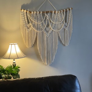 Puede incluir: Un gran tapiz de macram&eacute; blanco roto. La pieza est&aacute; suspendida de una varilla de madera natural y presenta intrincados patrones anudados y largas y fluidas flecos. Decoraci&oacute;n de pared.