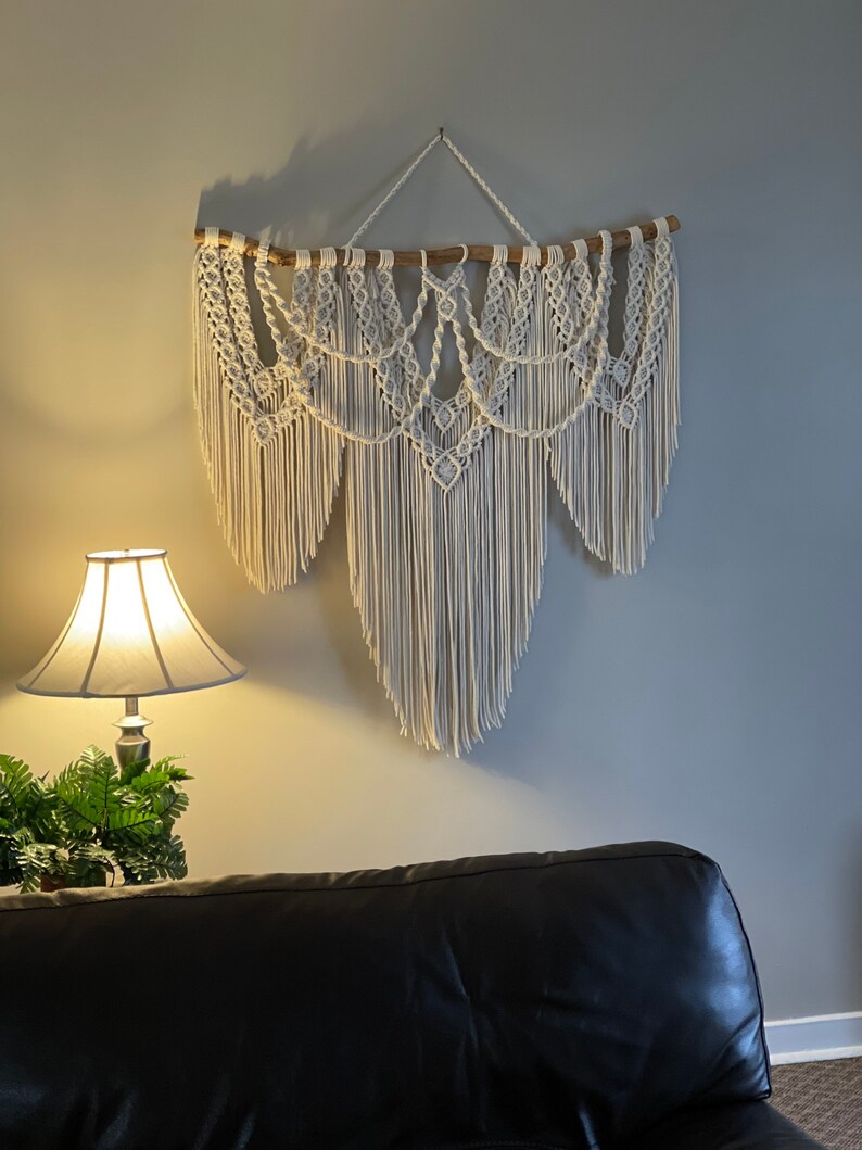 Puede incluir: Un gran tapiz de macram&eacute; blanco roto. La pieza est&aacute; suspendida de una varilla de madera natural y presenta intrincados patrones anudados y largas y fluidas flecos. Decoraci&oacute;n de pared.