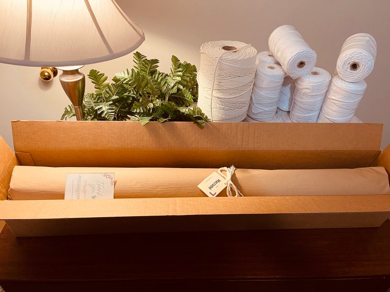 Puede incluir: Una caja de cart&oacute;n llena de suministros para manualidades. En el interior hay varias bobinas de cord&oacute;n de algod&oacute;n blanco, rollos de papel envuelto y una tarjeta peque&ntilde;a. Se ven plantas verdes con forma de helecho.