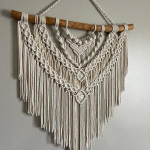 Peut inclure: Suspension murale en macramé crème avec un motif géométrique. La pièce est suspendue à une branche en bois naturel et présente des motifs de nœuds complexes. Le bas du macramé a une frange. Cette décoration murale artisanale ajoute une touche bohème.