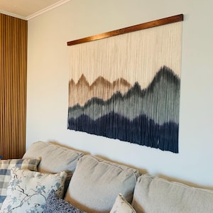 May include: A large wall hanging with a mountain design. The top portion is cream-colored, transitioning to shades of brown, gray, and navy blue at the bottom. It is suspended from a wooden rod. The wall hanging is above a gray sofa with decorative pillows.