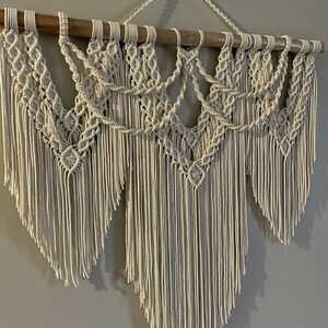 Puede incluir: Colgante de pared de macram&eacute; color crema. El dise&ntilde;o presenta intrincados patrones anudados y largas flecos, suspendidos de un list&oacute;n de madera natural. La decoraci&oacute;n de la pared es de estilo bohemio, agregando textura e inter&eacute;s visual.