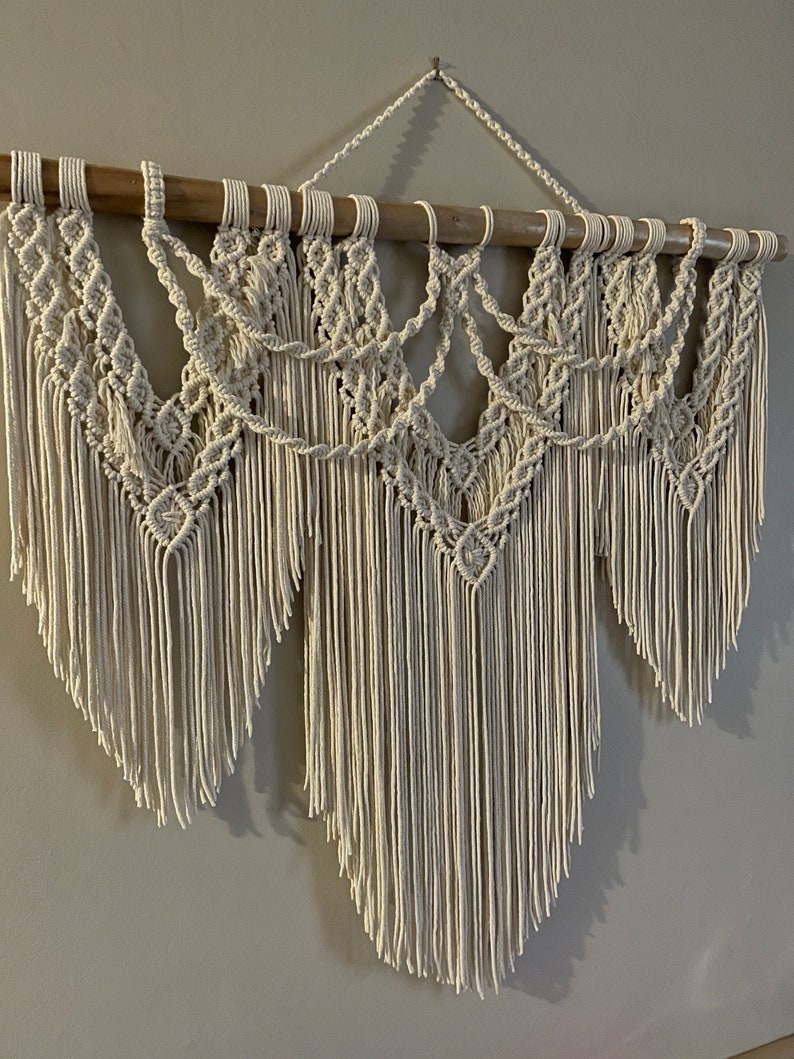 Puede incluir: Colgante de pared de macram&eacute; color crema. El dise&ntilde;o presenta intrincados patrones anudados y largas flecos, suspendidos de un list&oacute;n de madera natural. La decoraci&oacute;n de la pared es de estilo bohemio, agregando textura e inter&eacute;s visual.