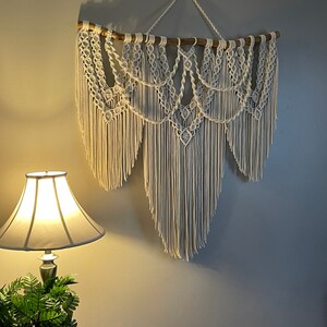 Puede incluir: Un gran tapiz de macram&eacute; blanco roto. La pieza est&aacute; suspendida de una rama de madera natural y presenta intrincados patrones anudados y largas y fluidas flecos. El tapiz est&aacute; montado en una pared gris.