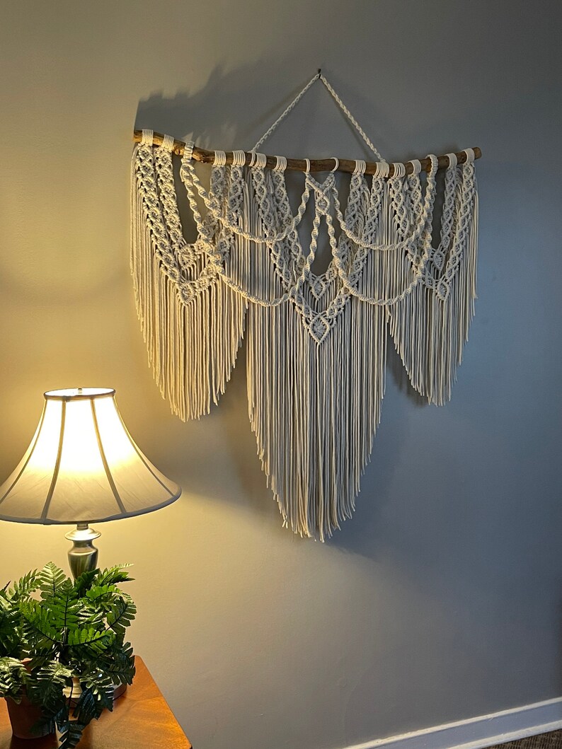Puede incluir: Un gran tapiz de macram&eacute; blanco roto. La pieza est&aacute; suspendida de una rama de madera natural y presenta intrincados patrones anudados y largas y fluidas flecos. El tapiz est&aacute; montado en una pared gris.
