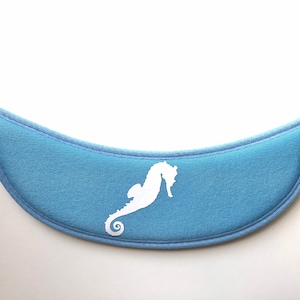 May include: A light blue visor brim featuring a white seahorse silhouette centered on the front. The curved accessory has a textured fabric surface with stitched edges, providing a clean and simple design for headwear applications.
