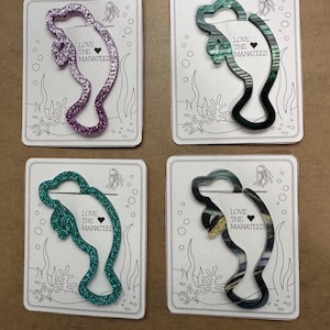 May include: Four manatee-shaped accessories in various colors: glittery purple, green, teal, and a marbled pattern. Each is on a white card with an underwater scene and the text "LOVE THE MANATEES".