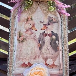 Op de afbeelding: Decoratief houten bord met een huwelijksthema-illustratie van twee eenden. Het bord is versierd met een jute strik, lavendel takjes en een grote perzikroos. Het frame is afgewerkt met kant, ongeveer 25 cm hoog.