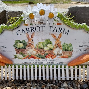 May include: A white wooden sign with a green border and a scalloped edge. The sign says "Locally Grown Carrot Farm" and features two bunnies holding carrots, surrounded by vegetables. The sign is decorated with white daisies and green leaves. The sign is sitting on a white picket fence.