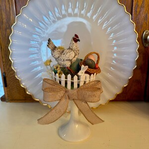 May include: A white ceramic pedestal with a brown burlap bow. Atop the pedestal is a white picket fence with a white rabbit, a brown basket with eggs, and a white chicken.