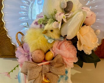 May include: A ceramic Easter train with a yellow chick, pink and white flowers, and speckled Easter eggs. The train is decorated with blue and white flowers and has a brown burlap bow.