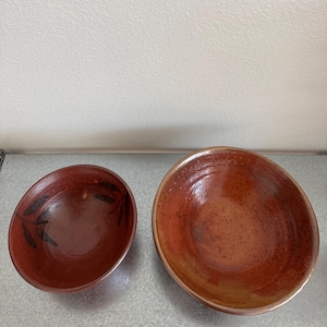 以下が含まれることがあります： 光沢のある仕上げの丸い陶器のボウル2つ。左側の小さいボウルは濃い赤茶色で、黒い葉のような模様があります。右側の大きいボウルは、より明るいまだら模様の赤オレンジ色で、内側の表面は滑らかで光を反射しています。