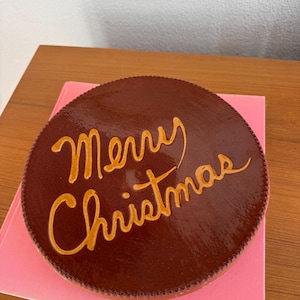 May include: A round, dark brown, glossy decorative disc with a textured, beaded edge. The words Merry Christmas are written across the center in a cursive, light orange, raised script font.