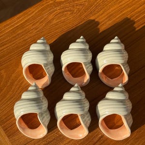 May include: A set of six ceramic napkin rings shaped like spiral seashells. Each piece features a glossy, off-white exterior with a smooth, ribbed texture and a contrasting soft peach-colored interior opening, arranged in two rows of three.