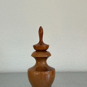 May include: A small, turned wooden vessel with a smooth, polished finish and visible grain patterns. The object features a wide, rounded base that tapers into a narrow neck, topped with a distinct, tiered lid that ends in a pointed finial.