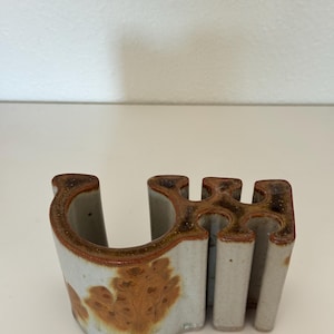 May include: A ceramic desk organizer with a light gray base and a brown, mottled glaze on the top edges and side. The object features a large curved opening on one side and three vertical, cylindrical slots on the other side for holding items.