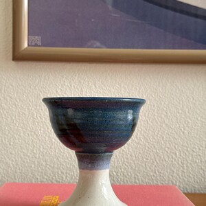 May include: A ceramic chalice featuring a deep blue and purple glazed bowl that transitions into a lighter lavender band, supported by a solid off-white pedestal base. The vessel has a wide, flared rim and a smooth, glossy finish throughout.