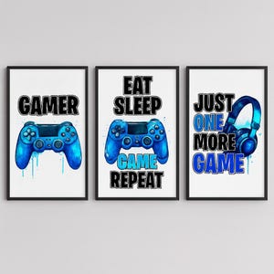 May include: Three framed prints with a gaming theme. Each print has a black frame and a white background. One print says "GAMER" with a blue controller graphic. Another says "EAT SLEEP GAME REPEAT" with a blue controller. The third says "JUST ONE MORE GAME" with a blue headset.