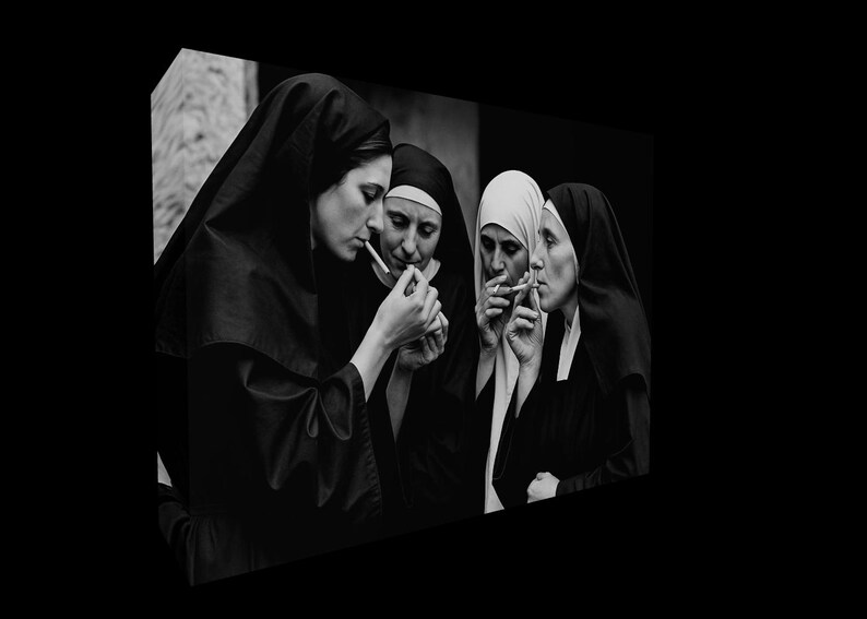 May include: A monochrome image showing four nuns in their habits, smoking cigarettes. One nun is using a lighter to light a cigarette for another. The image uses light and shadow to create a striking contrast.