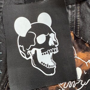 May include: Close-up of a black denim jacket with patches. One patch features a white skull with mouse ears, another says "REBEL REBEL" in white, and a third has a faded design. The jacket has a zipper.