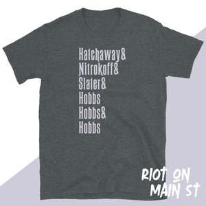 May include: A dark gray t-shirt with white text that reads "Hathaway & Nitrokoff & Slater & Hobbs & Hobbs & Hobbs" and "Riot on Main St" in the bottom right corner.