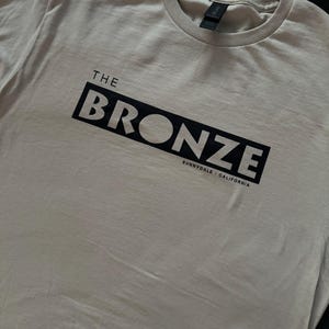May include: A light gray t-shirt with a black box that says "THE BRONZE" and "SUNNYDALE, CALIFORNIA" in white text.