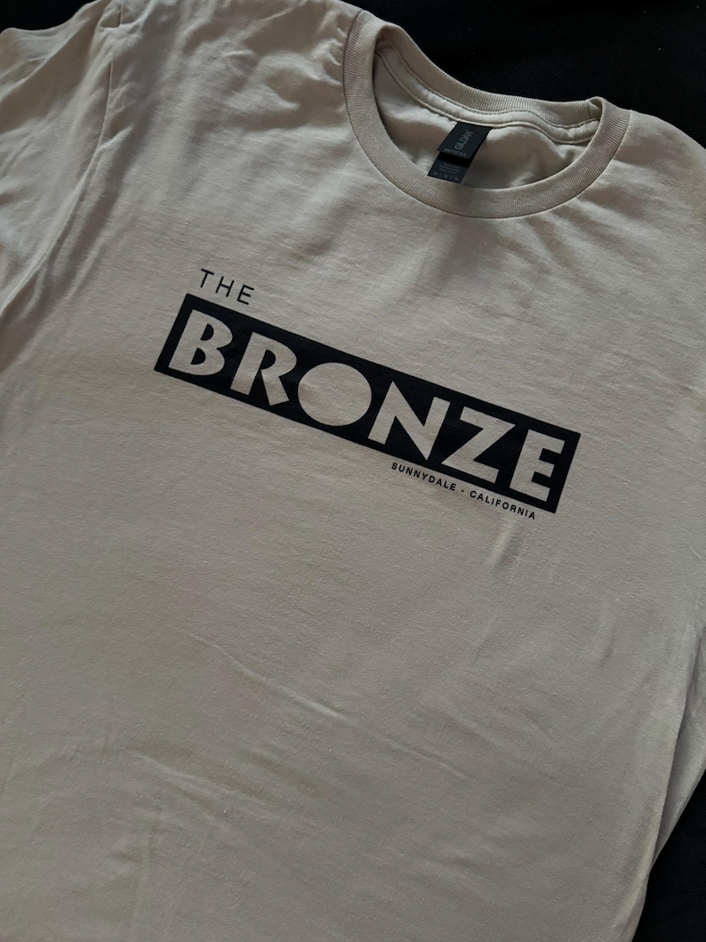 May include: A light gray t-shirt with a black box that says "THE BRONZE" and "SUNNYDALE, CALIFORNIA" in white text.