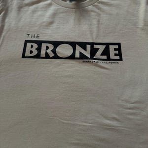 May include: A beige t-shirt with a black graphic that says "THE BRONZE SUNNYDALE - CALIFORNIA"