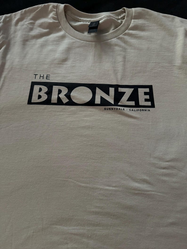 May include: A beige t-shirt with a black graphic that says "THE BRONZE SUNNYDALE - CALIFORNIA"