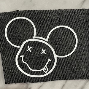 May include: A black fabric patch with a white outline of a smiling Mickey Mouse head with X's for eyes and a tongue sticking out.