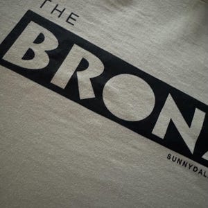 May include: A white t-shirt with black text that reads "THE BRONX" and "SUNNYDALE" in a bold, blocky font.