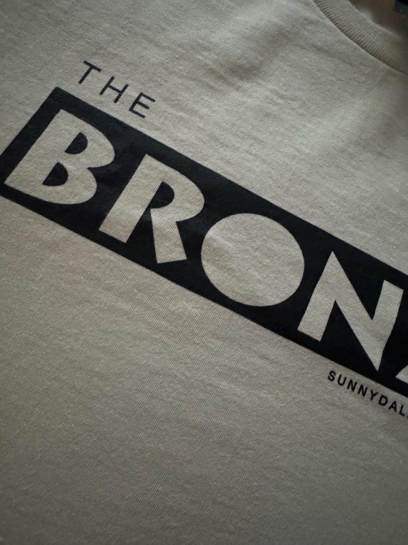 May include: A white t-shirt with black text that reads "THE BRONX" and "SUNNYDALE" in a bold, blocky font.